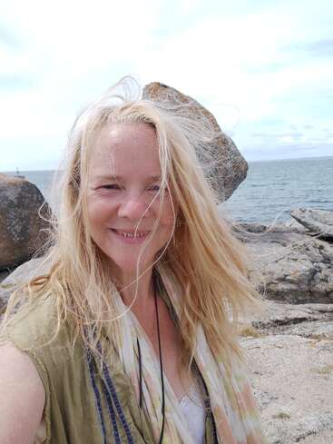 Una mujer rubia sonríe a la cámara en una playa rocosa. Lleva el pelo al viento, el cielo está nublado y el mar está detrás de ella.