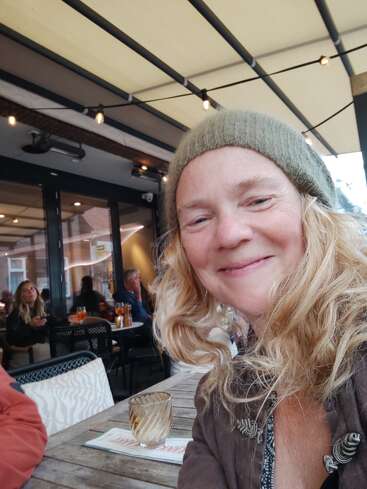 Una mujer con el pelo rubio y un gorro verde sonríe a la cámara sentada en un café al aire libre. Al fondo se ve a otras personas socializando.