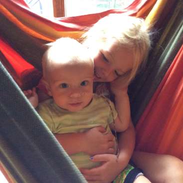 Das Bild zeigt ein junges Mädchen mit blonden Haaren, das ein Baby in einer Hängematte hält, wobei das Gesicht des Mädchens nahe am Kopf des Babys ist.