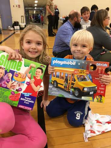 Das Bild zeigt zwei Kinder, die auf dem Boden einer Turnhalle sitzen und jeweils eine Spielzeugkiste in der Hand halten, mit einer Menschenmenge im Hintergrund, wahrscheinlich bei einer Schulveranstaltung oder einer Versammlung.