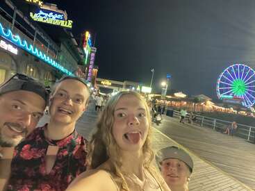 A happy family takes a fun selfie on a lively boardwalk at night, with bright neon lights and a colorful Ferris wheel glowing in the background.