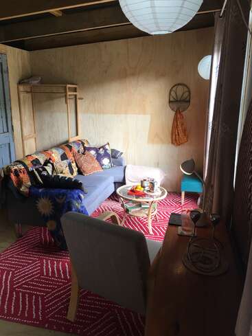 Un acogedor salón cuenta con un sofá azul con colchas de colores, una alfombra con motivos rojos, una mesa de centro redonda, muebles sencillos de madera y luz natural que entra por una ventana.