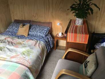 Se trata de un dormitorio acogedor con una cama bien hecha con ropa de cama estampada, una mesita de noche con una lámpara y una taza, un sillón, una cómoda con una planta y una iluminación tenue.