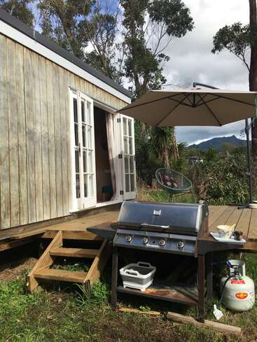 Una cabaña rústica de madera con puertas acristaladas que dan a una terraza. Una barbacoa, una sombrilla, una bombona de propano y una silla de exterior crean un ambiente acogedor y relajado.