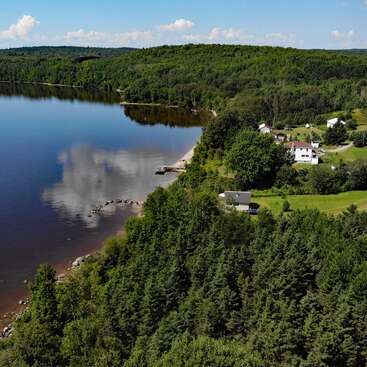 A serene lakeside scene features calm water reflecting clouds, dense green forest, and scattered houses. The coastline and lush landscape create a peaceful, idyllic rural setting.