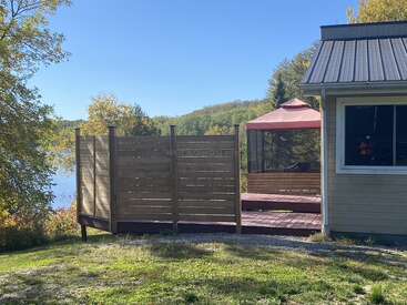 Uma área aconchegante à beira do lago com uma cerca de madeira para privacidade, um gazebo com tela e uma casa. Vedeção verdejante ao redor, com um lago e um céu azul ensolarado.