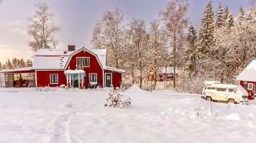 Ein charmantes rotes Haus steht in einer verschneiten Landschaft, umgeben von blattlosen Bäumen, ein weißer Lieferwagen parkt in der Nähe und das Sonnenlicht beleuchtet die winterliche, ruhige Waldszene.