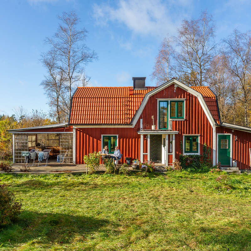 Una encantadora casa de madera roja con un empinado tejado naranja se asienta en un patio cubierto de hierba. Dos personas se relajan al aire libre bajo el cielo azul, disfrutando de la naturaleza y el sol.
