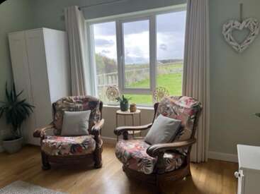 Two cozy armchairs with floral patterns sit by a large window, overlooking green fields. Decor includes plants, a white wardrobe, and a driftwood heart wall hanging.