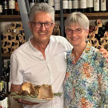 An older smiling couple stands in front of wine shelves. The man holds a plate of mushrooms with herbs. Both wear glasses and patterned shirts, appearing happy.