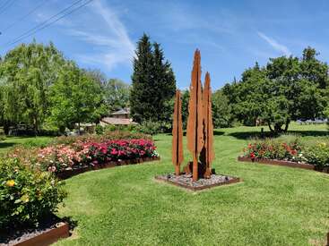 Dieses Bild zeigt einen wunderschönen Garten mit bunten Blumenbeeten, grünem Gras, großen Bäumen und einer hohen, modernen rostfarbenen Metallskulptur in der Mitte.