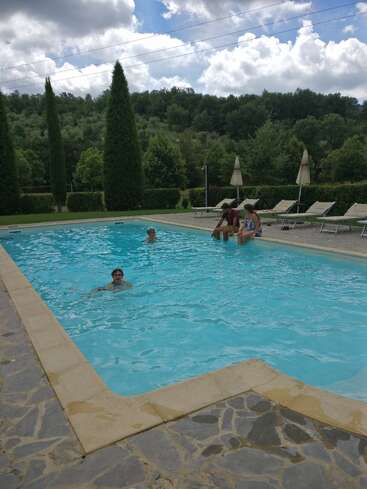 Ein ruhiger Außenpool, umgeben von Grün und hohen Bäumen. Drei Personen sitzen am Rand, während eine Person schwimmt. Poolstühle und Sonnenschirme säumen die Seite.