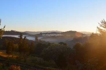 The image depicts a serene landscape with rolling hills, trees, and a misty valley, set against a clear blue sky with the sun shining brightly.