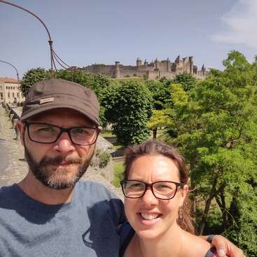 A smiling couple wearing glasses stands outdoors, taking a selfie. Behind them, lush green trees and a magnificent medieval castle rise against the bright blue sky.