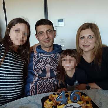 A family of four poses together, smiling around a table with a birthday cake displaying the number 46. The children's faces are pixelated for privacy.