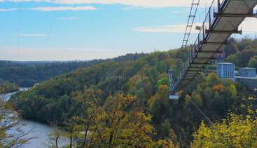 A suspension footbridge stretches high above a river, surrounded by dense, colorful forest. The sky is partly cloudy, creating a peaceful, scenic natural landscape.