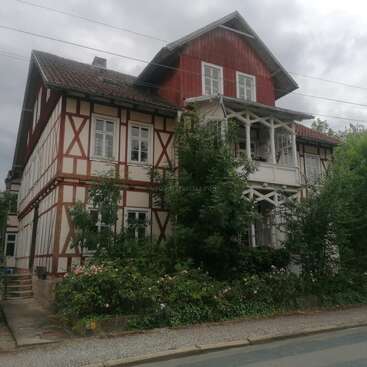 A charming, traditional half-timbered house with red and white colors, lush greenery in front, a small balcony, and a steep, tiled roof under cloudy skies.
