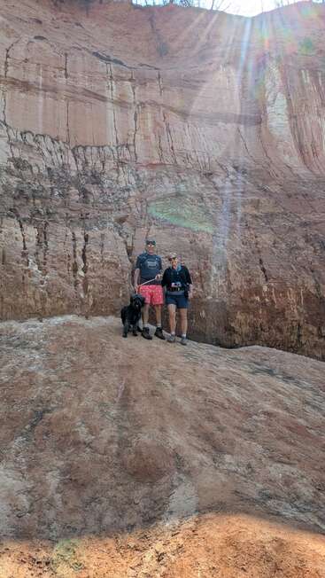 Zwei Menschen und ein schwarzer Hund stehen auf felsigem Gelände vor einer hohen, geschichteten Klippe, gebadet in Sonnenlicht, das von der rechten oberen Ecke herabströmt.