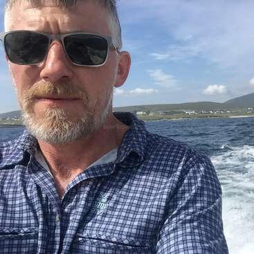 A imagem mostra um homem com cabelos grisalhos e barba, usando óculos escuros, em frente a um corpo d'água com uma paisagem montanhosa ao fundo.