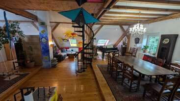 This spacious loft features a spiral staircase, wooden beams, dining area, lounge with colorful wall art, piano, plants, grandfather clock, chandelier, and large windows. Cozy atmosphere.