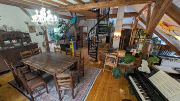 This image shows a cozy attic living space with wooden beams, a spiral staircase, dining table, chandelier, plants, piano, books, and colorful, artistic decor.