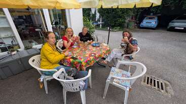 Four people sit around a colorful table outdoors, enjoying food and drinks under yellow umbrellas. A small dog rests on a chair. Cars are parked nearby.