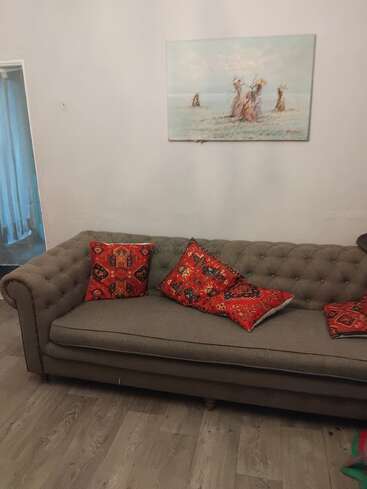 Un sofá gris empenachado se apoya en una pared clara, decorada con cojines rojos estampados. Encima, un cuadro en tonos pastel muestra figuras, mientras que el suelo de madera completa la acogedora escena.