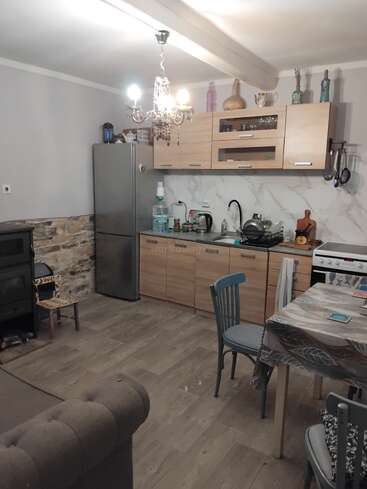 Una acogedora cocina con armarios de madera, frigorífico de acero inoxidable, detalles de piedra, lámpara de araña, una pequeña mesa de comedor, fogones y varios utensilios de cocina ordenados.