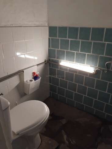 Esta imagen muestra un pequeño cuarto de baño con un inodoro, paredes alicatadas en azul y blanco, una barra de sujeción, una cesta de pared con artículos de aseo y un fluorescente encendido.