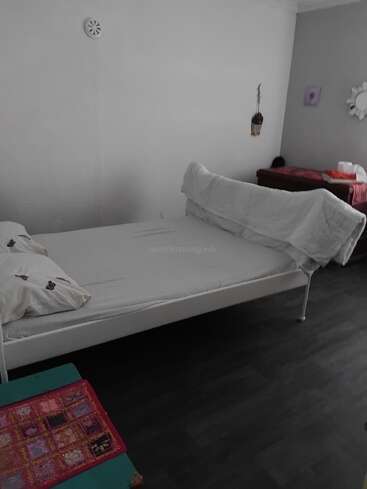 Un dormitorio sencillo y acogedor con una cama blanca, almohadas estampadas, pequeños adornos en la pared, una mesa cubierta de rojo, suelo oscuro y una colorida tela bordada sobre un banco verde.