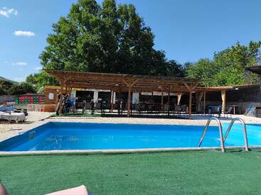Una piscina exterior transparente con escalones metálicos, rodeada de césped artificial verde, se asienta frente a una zona de patio con sombra de madera y frondosos árboles verdes.