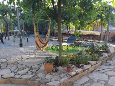 Ein friedlicher Garten mit einer farbenfrohen Hängematte, steinernen Wegen, Topfpflanzen, Liegestühlen auf grünem Gras und üppig überhängenden Weinreben bietet einen entspannenden Rückzugsort.
