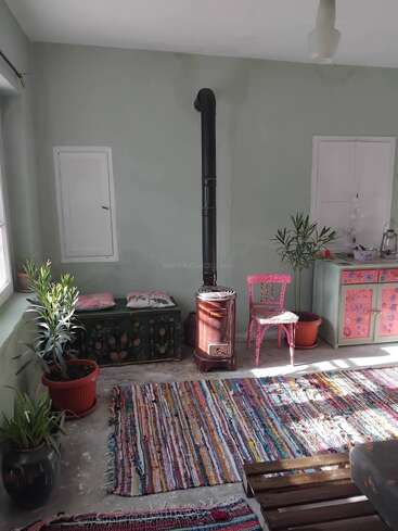 Um quarto aconchegante com um fogão a lenha, tapetes coloridos, vasos de plantas, um baú pintado, uma cadeira rosa e um armário com motivos florais iluminado pela luz do sol.