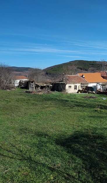 Une scène champêtre met en scène une vieille maison au toit rouge, des remises et une grande cour herbeuse. Des arbres dépouillés et des collines verdoyantes ondulantes se dessinent sous un ciel d'un bleu éclatant.