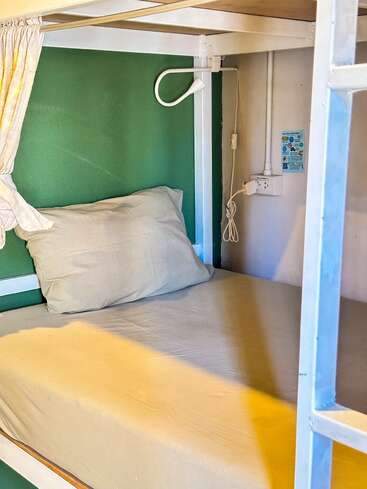 This image shows a neatly made bunk bed with beige linens, a pillow, a reading light, power outlet, and a green wall. A curtain offers some privacy.