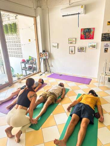 Three people are lying on yoga mats in a sunlit room, practicing relaxation while an instructor gently assists. Art, plants, and natural light create a peaceful atmosphere.