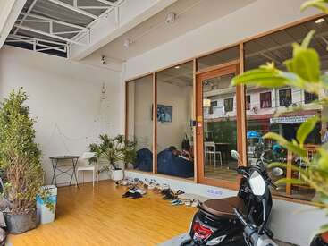 A cozy café entrance with wooden flooring, potted plants, and several pairs of shoes outside. Glass doors reveal a relaxed interior. A motor scooter is parked nearby.