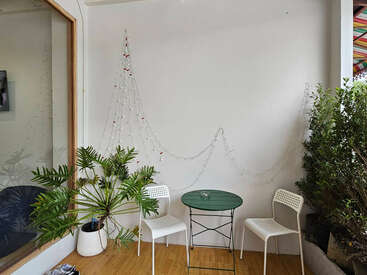 A cozy corner with two white chairs and a green round table, decorated with a potted plant, fairy lights on the wall, and wooden flooring.