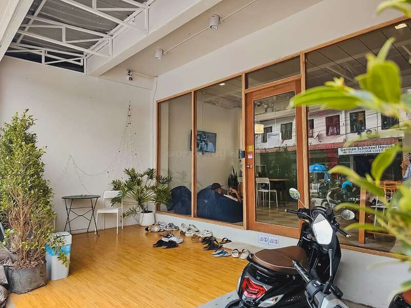 A cozy café entrance with wooden flooring, potted plants, and several pairs of shoes outside. Glass doors reveal a relaxed interior. A motor scooter is parked nearby.