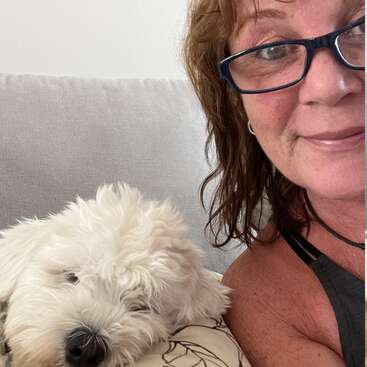 A woman with glasses smiles next to a fluffy white dog resting on a couch. The scene looks cozy and relaxed, capturing a peaceful moment together.