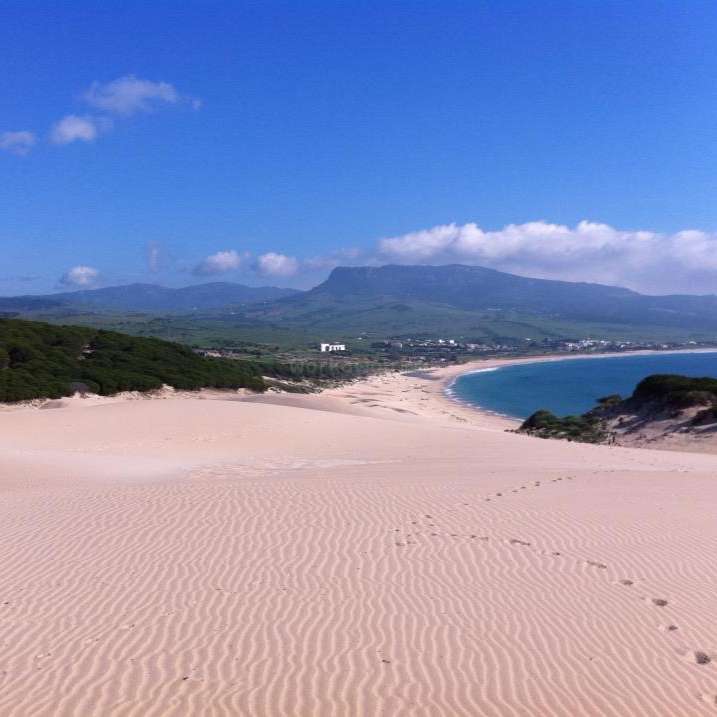 Das Bild zeigt eine ruhige Strandszene mit einer weiten Sandfläche, einem ruhigen Gewässer und einer majestätischen Bergkette im Hintergrund unter einem klaren blauen Himmel.