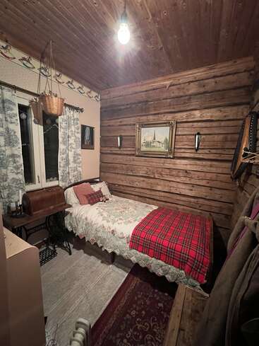 A cozy, rustic bedroom with wooden walls and ceiling, single bed, plaid and patchwork bedding, vintage sewing machine desk, hanging plant, curtains, and framed painting.
