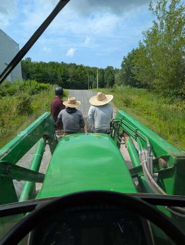 Three people, two wearing straw hats, sit on the green front loader of a tractor, traveling down a rural path surrounded by trees and greenery under blue sky.