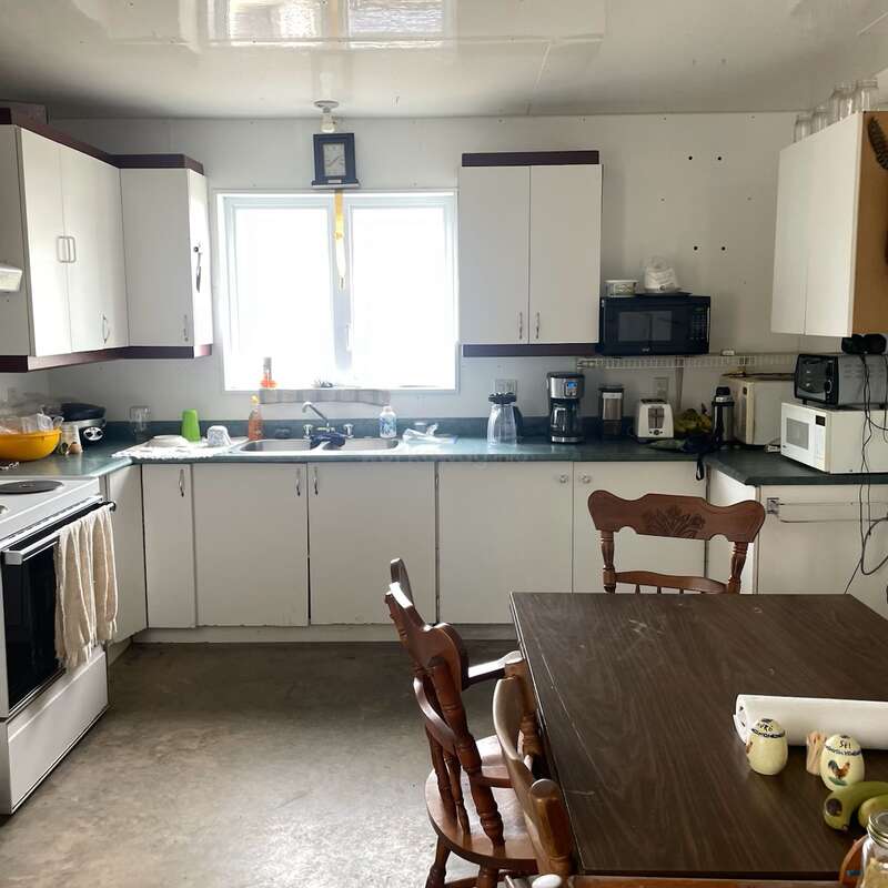 This kitchen features white cabinets, a green countertop, appliances, and a wooden dining table with chairs. Natural light comes through the window above the sink.