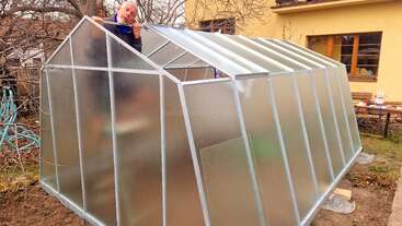 Una persona está montando un invernadero translúcido en un patio trasero. La estructura tiene un armazón metálico y paneles. Se ven árboles, una casa y una mesa cerca.