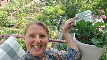 Una mujer sonriente se hace una alegre selfie en un balcón, señalando unas flores de color rosa intenso en una maceta, con un frondoso jardín verde visible al fondo.