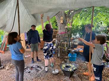 Five people are outdoors under a tarp. Some cook on an open fire, while others adjust the tarp. The atmosphere is casual and communal, surrounded by trees.