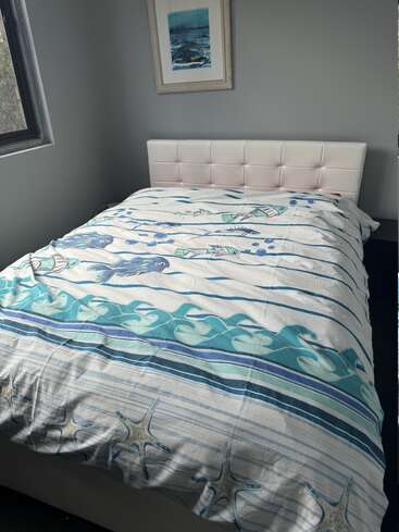 Un lit bien fait avec un couvre-lit sur le thème de la mer, avec des poissons, des vagues et des étoiles de mer. La chambre a des murs gris, une fenêtre et un tableau encadré représentant l'océan.