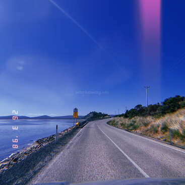 Une route côtière sinueuse suit le littoral sous un ciel d'un bleu éclatant. Des panneaux de signalisation, des collines herbeuses, de l'eau et des montagnes lointaines créent un paysage paisible et pittoresque.