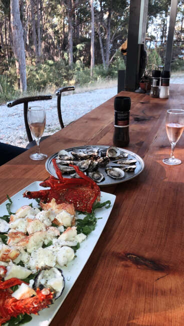 Une table rustique en bois en plein air présente un festin de fruits de mer avec du homard et des huîtres, accompagné de verres de vin, de moulins à poivre et entouré d'un paysage forestier luxuriant.